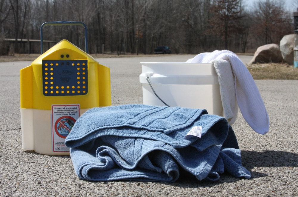 A folded blue towel is on the ground in front of a yellow and white bait bucket and a white bucket with a white towel draped over the rim. These items are all on concrete pavement and there are bare trees in the background.