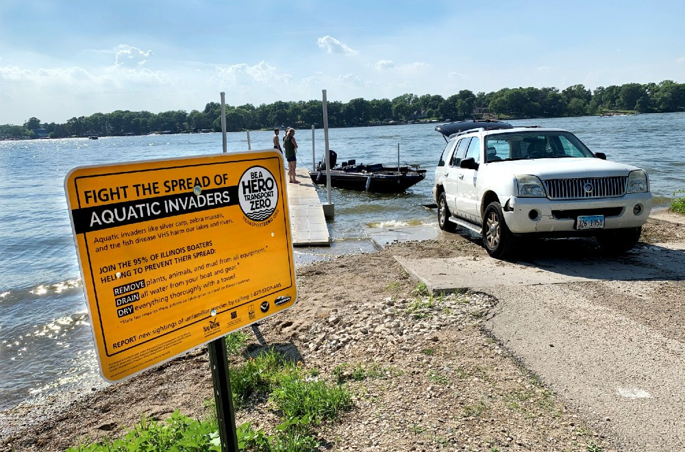 A white car backs into the water at a boat launch at a large lake or river. There is a boat in the water behind the car and two people on a dock next to the boat. In the foreground is an orange sign with the text 