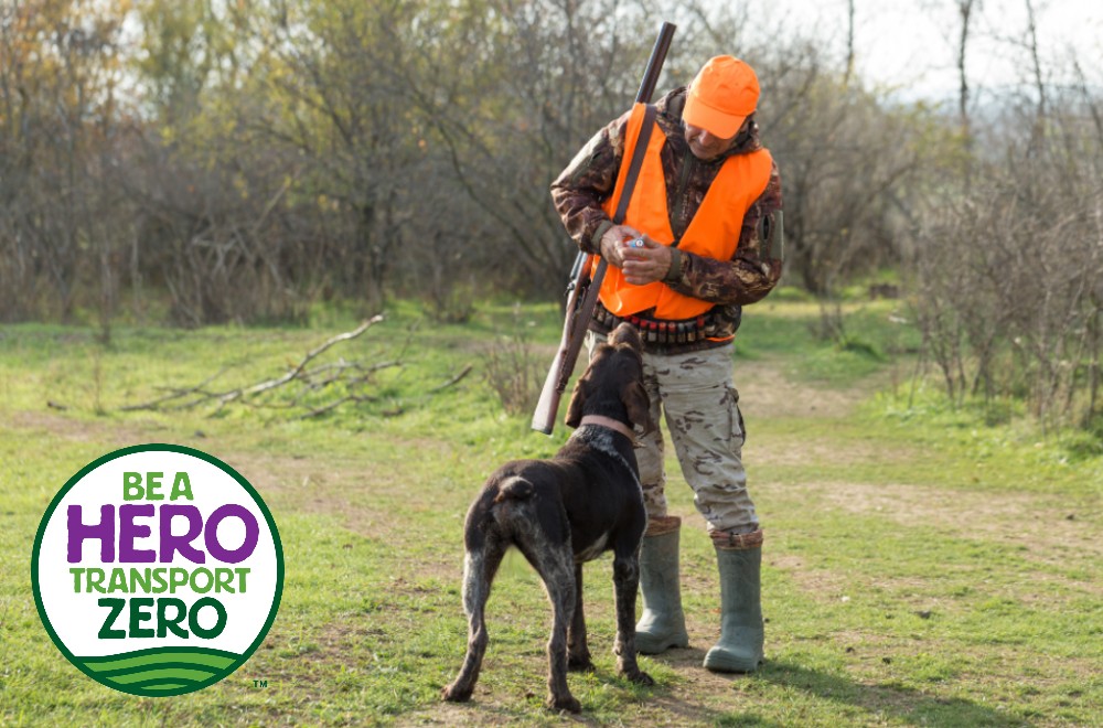 A hunter in an orange vest and hat with a gun slung over his shoulder stands looking down at a brown dog in front of him. They are in grass with bare trees and shrubs behind them. Superimposed on the bottom left corner is a circular logo with the text 