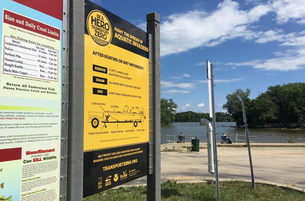 Two informational signs in the parking lot of a boat launch. One sign has information about fish, and the other sign has information about how to clean a boat of aquatic invasive species. There are people sitting in chairs in the background near the water.