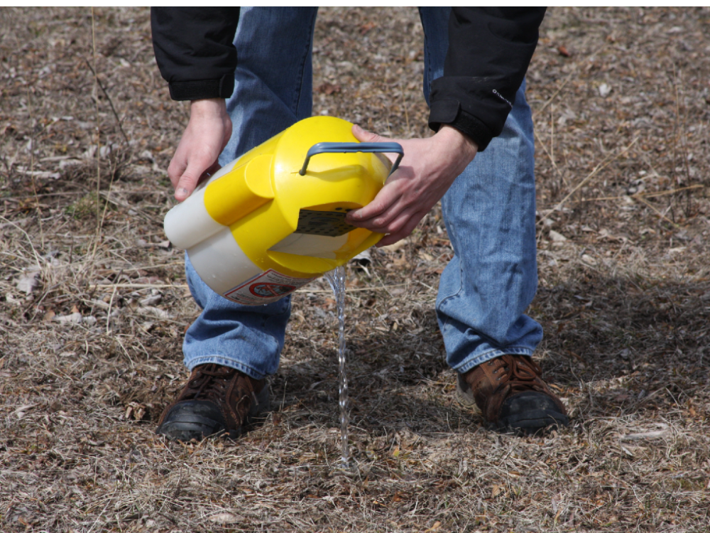 Close-up of a person holding a yellow bait bucket pouring the water out into the brown grass. They are wearing a black jacket, blue jeans and brown shoes.