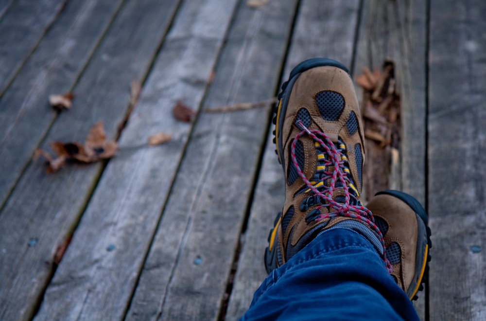 Close-up of brown hiking boots with red laces. The person wearing them is crossing their legs and they are also wearing blue pants. Their feet are resting on a wooden boardwalk and there are some fallen leaves in the background.